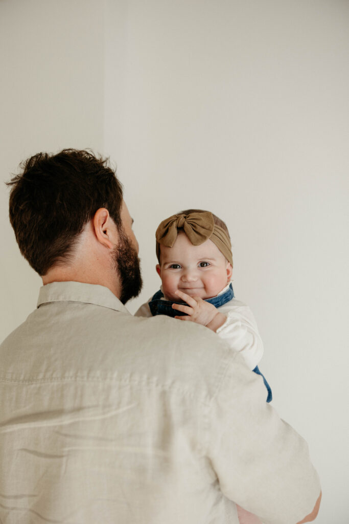 baby peeking out smiling behind dad's shoulder during a studio photoshoot with soft natural lighting